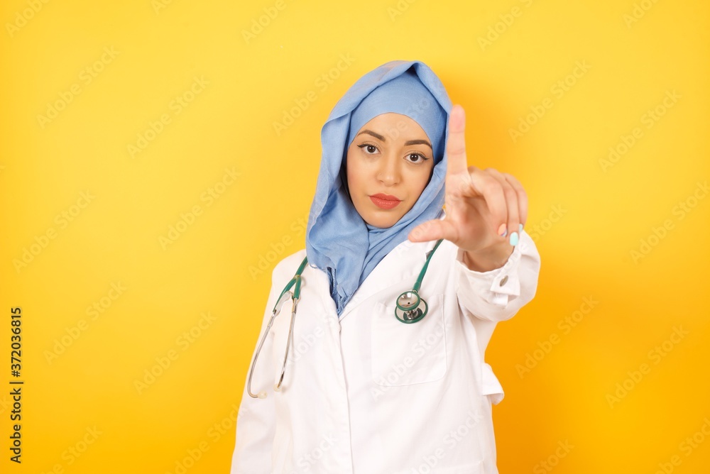 Φωτογραφία Young arab doctor woman wearing medical uniform standing over yellow background  making fun of people with fingers on forehead doing loser gesture mocking and insulting