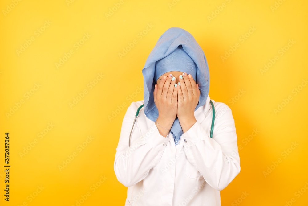 Φωτογραφία Young arab doctor woman wearing medical uniform standing over yellow background  covering her face with her hands, being devastated and crying