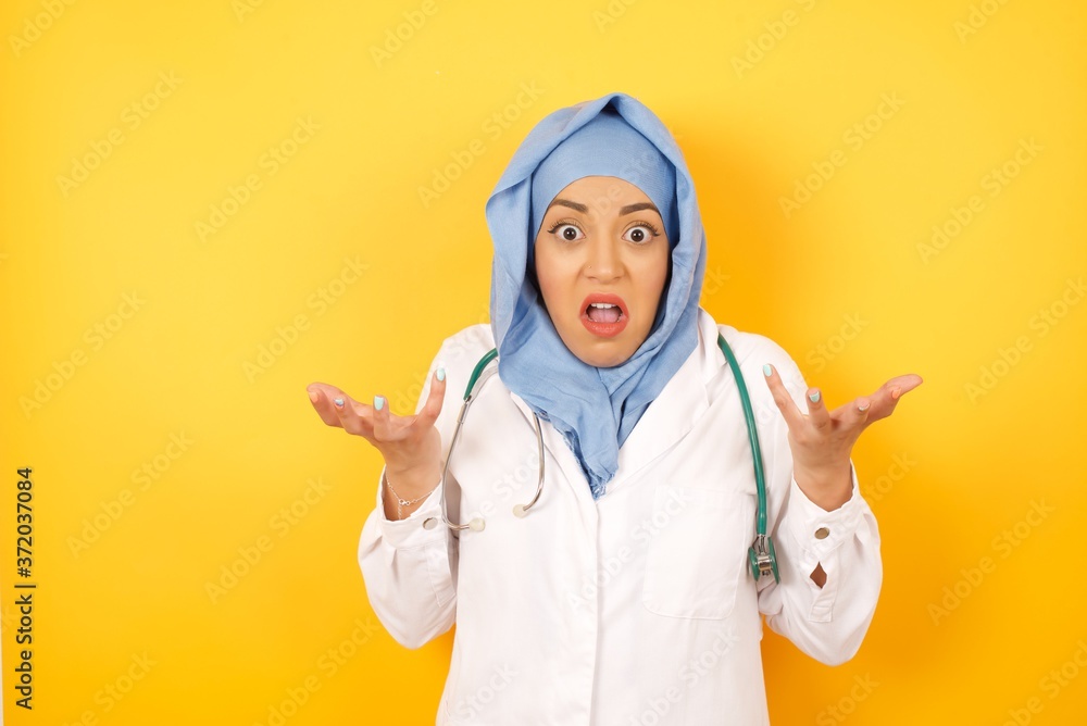 Φωτογραφία Frustrated Young arab doctor woman wearing medical uniform standing over yellow background  puzzled and hesitant, shrugs shoulders in bewilderment, keeps mouth widely opened, doesn't know what to do