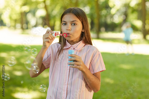 Waist up portrait of teenage girl blowing bubbles while standing in green park outdoors, copy space