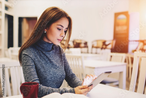 Young beautiful woman surfing internet websites for online shopping and browsing application on digital touch pad device sitting in wifi zone of stylish coffee shop interior with red cup of tea