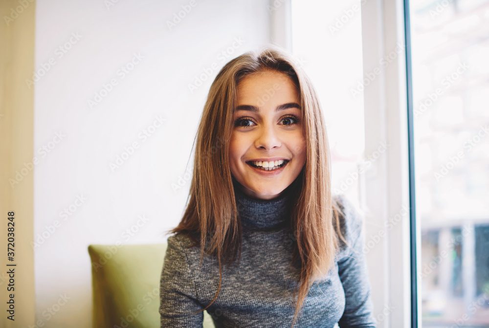 Half length portrait of beautiful cheerful woman dressed in casual wear looked at camera sitting in coworking space,attractive smiling hipster girl having relaxation time during work break indoors
