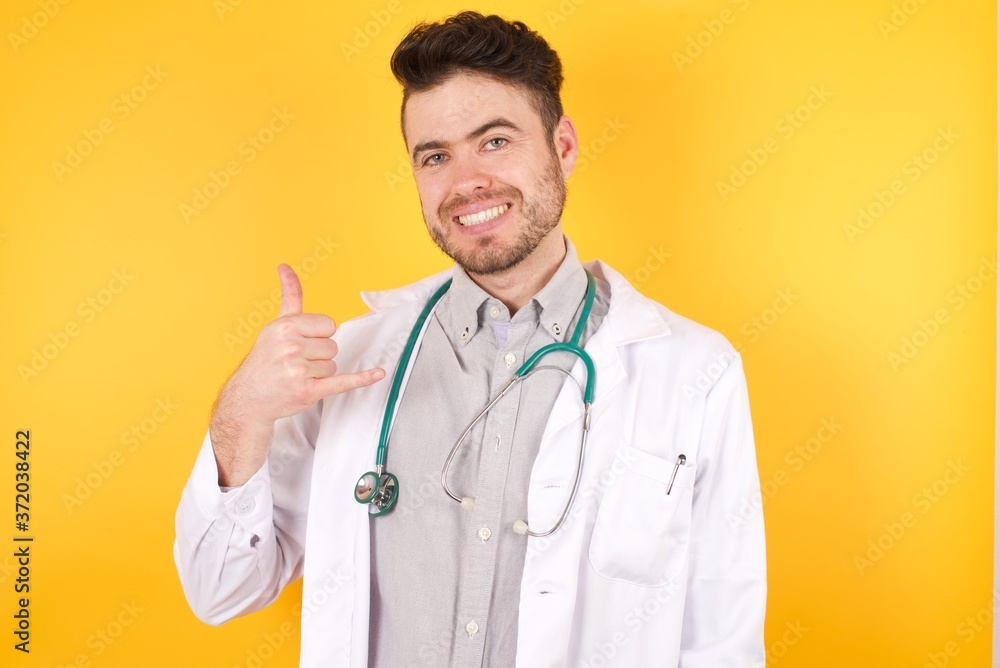 Young handsome Caucasian man wearing medical uniform making call gesture with hands. Lovely attractive man standing against white wall. Young handsome Caucasian man wearing medical uniform making call gesture with hands. Lovely attractive man standing against white wall.
