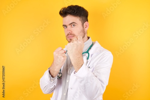 Young Caucasian doctor man wearing medical uniform  standing over isolated yellow background Ready to fight with fist defense gesture  angry and upset face  afraid of problem.