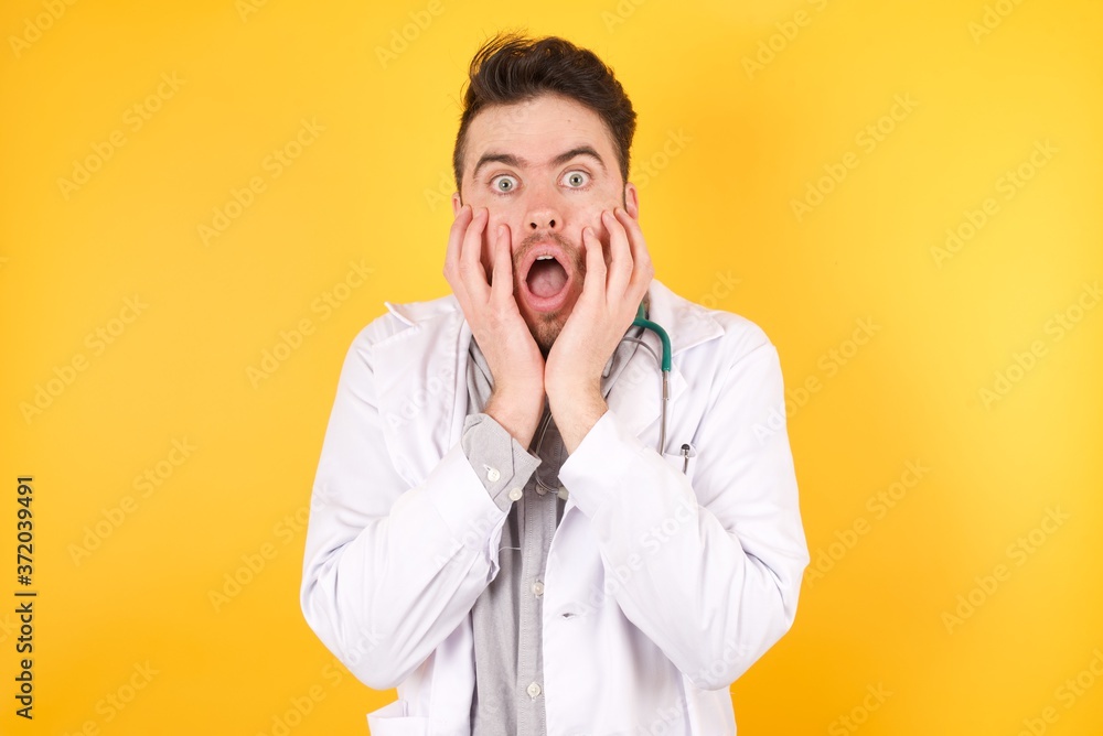 Stupefied young handsome Caucasian doctor man wearing medical uniform, expresses excitement and thrill, keeps jaw dropped, hands on cheeks, has eyes popped out.