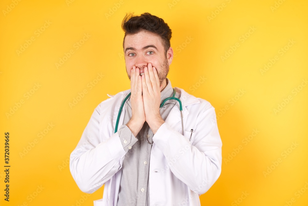 Young vivacious handsome young Doctor man wearing medical uniform giggles joyfully, covers mouth, has fun alone, stands against background or studio wall, has natural laughter, Young vivacious handsome young Doctor man wearing medical uniform giggles joyfully, covers mouth, has fun alone, stands against background or studio wall, has natural laughter,