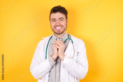 Positive adorable doctor man wearing medical uniform smiles happily, glad to receive pleasant news from his patient, keeps hands together. Medicine, health and emotions concept.