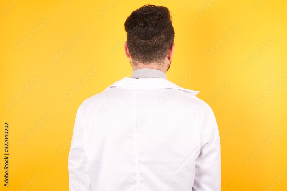 The back view of a young handsome doctor man wearing medical uniform, standing against yellow wall. Studio Shoot. The back view of a young handsome doctor man wearing medical uniform, standing against yellow wall. Studio Shoot.