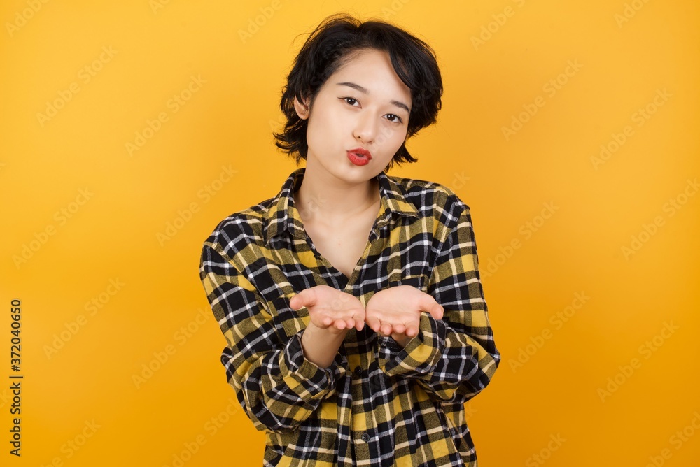 Young asian woman with short hair wearing plaid shirt standing over yellow background sending blow kiss with pout lips and holding palms to send air kiss. Young asian woman with short hair wearing plaid shirt standing over yellow background sending blow kiss with pout lips and holding palms to send air kiss.