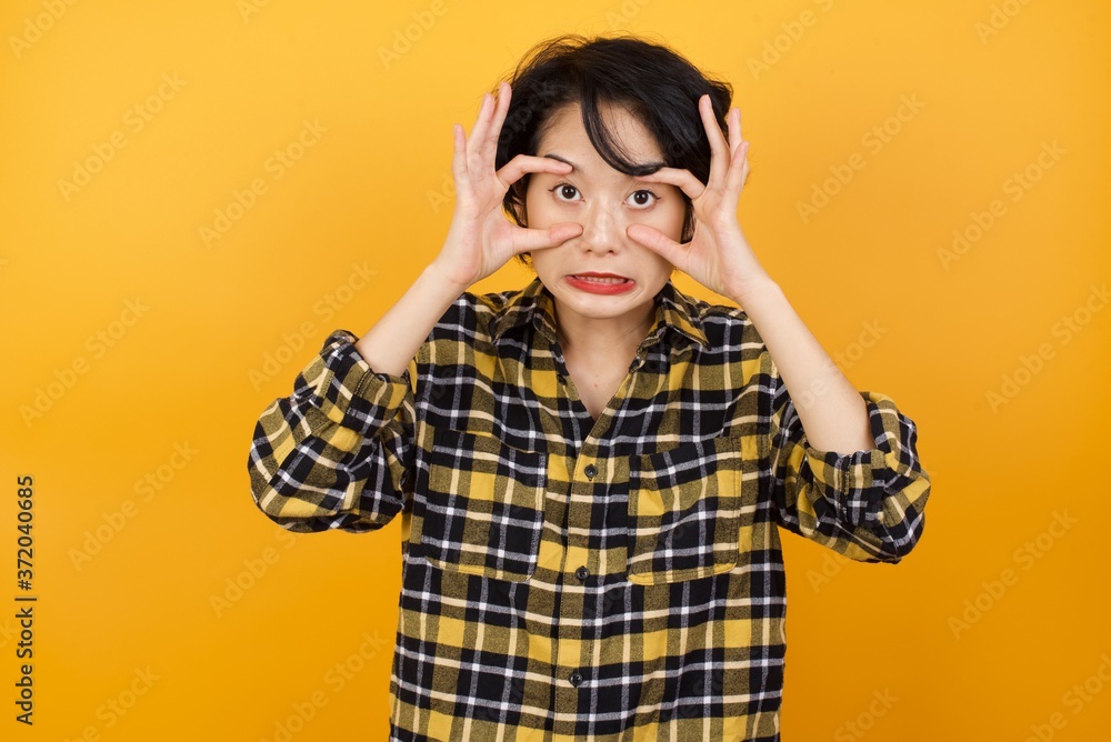 Young asian woman with short hair wearing plaid shirt standing over yellow background keeping eyes opened to find a success opportunity. Young asian woman with short hair wearing plaid shirt standing over yellow background keeping eyes opened to find a success opportunity.