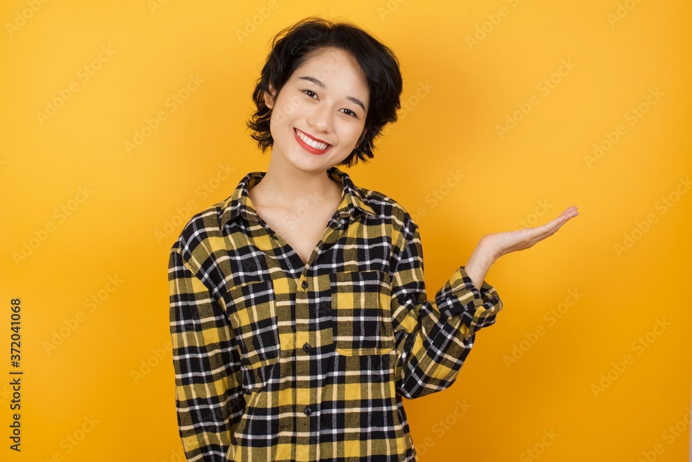 Young asian woman with short hair wearing plaid shirt standing over yellow background smiling cheerful presenting and pointing with palm of hand looking at the camera. Young asian woman with short hair wearing plaid shirt standing over yellow background smiling cheerful presenting and pointing with palm of hand looking at the camera.