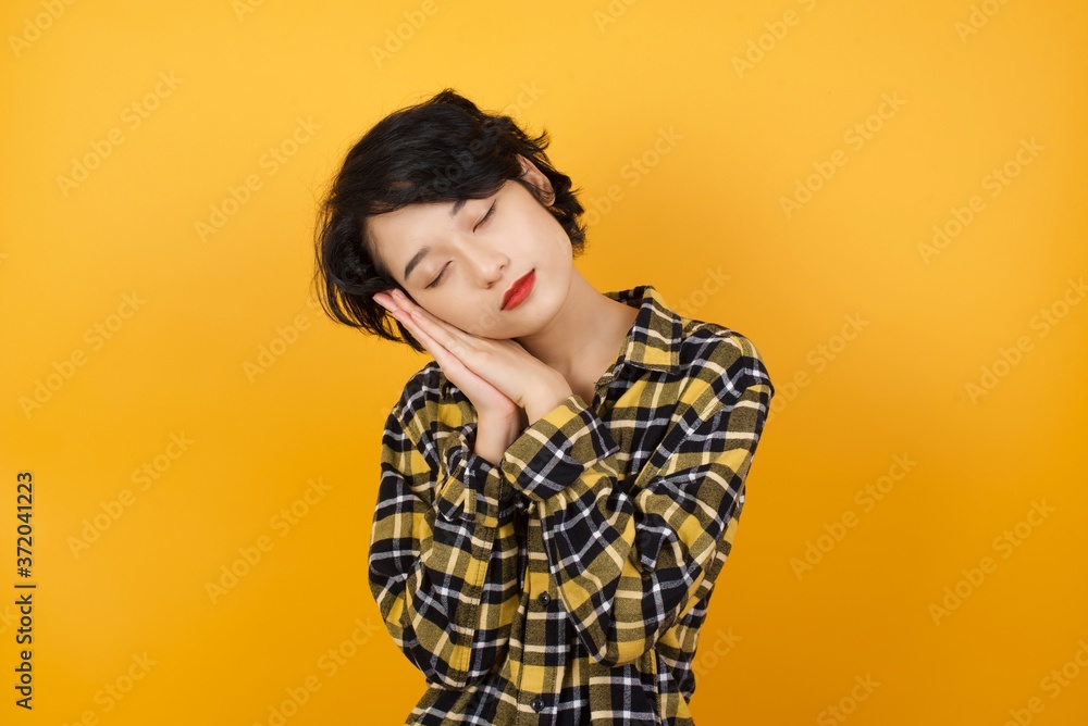 Young asian woman with short hair wearing plaid shirt standing over yellow background sleeping tired dreaming and posing with hands together while smiling with closed eyes. Young asian woman with short hair wearing plaid shirt standing over yellow background sleeping tired dreaming and posing with hands together while smiling with closed eyes.