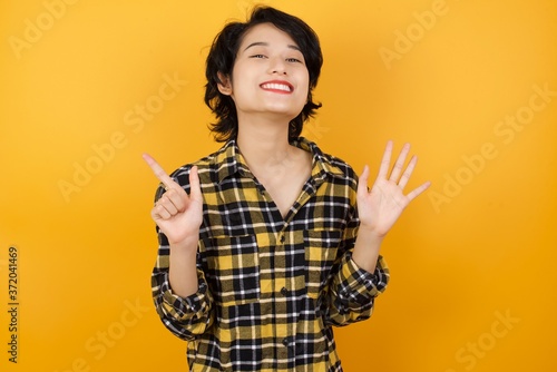 Young asian woman with short hair wearing plaid shirt standing over yellow background showing and pointing up with fingers number seven while smiling confident and happy.