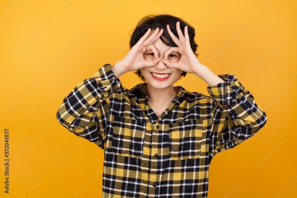 Young beautiful Asian woman wearing plaid shirt over yellow background doing ok gesture like binoculars sticking tongue out, eyes looking through fingers. Crazy expression. Young beautiful Asian woman wearing plaid shirt over yellow background doing ok gesture like binoculars sticking tongue out, eyes looking through fingers. Crazy expression.