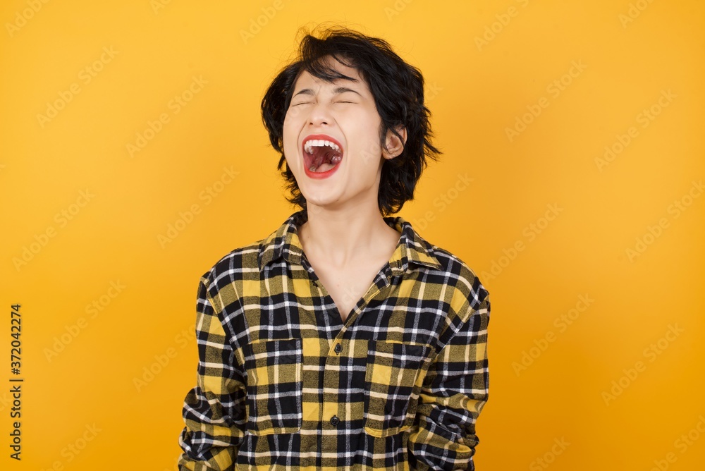 Young beautiful Asian woman wearing plaid shirt over yellow background angry and mad screaming frustrated and furious, shouting with anger. Rage and aggressive concept. Young beautiful Asian woman wearing plaid shirt over yellow background angry and mad screaming frustrated and furious, shouting with anger. Rage and aggressive concept.