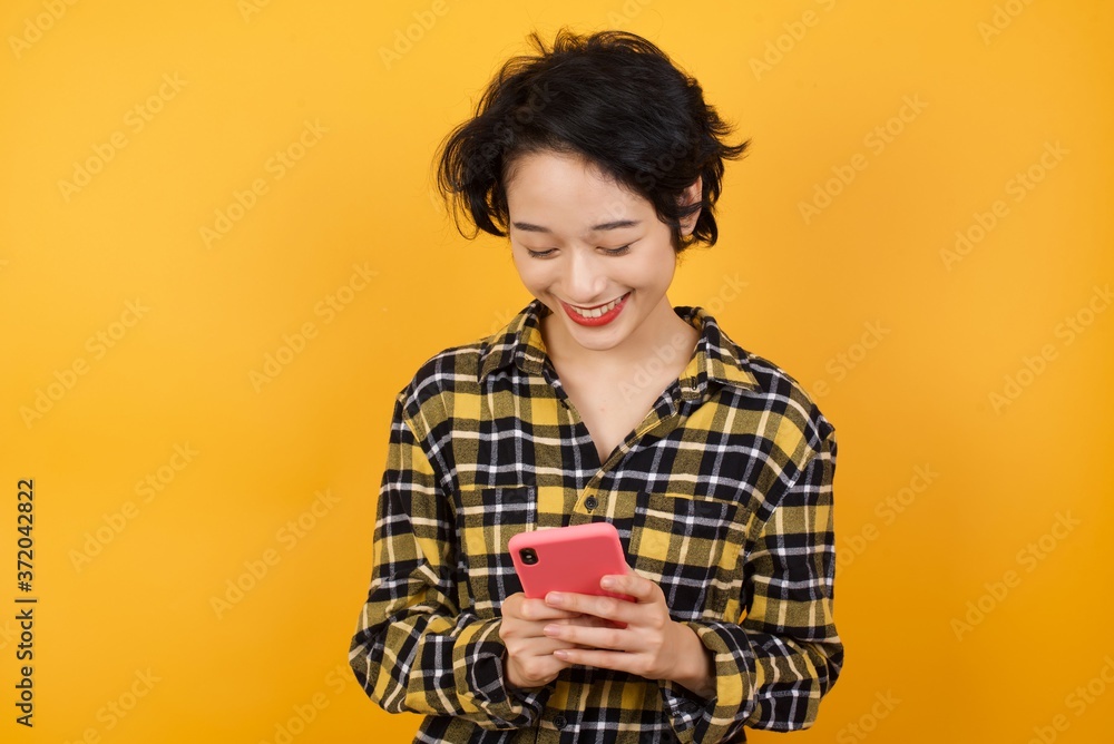 Young beautiful Asian woman wearing plaid shirt enjoys distant communication, uses mobile phone, surfs fast unlimited internet, has pleasant smile, dressed in red shirt, makes shopping online, 