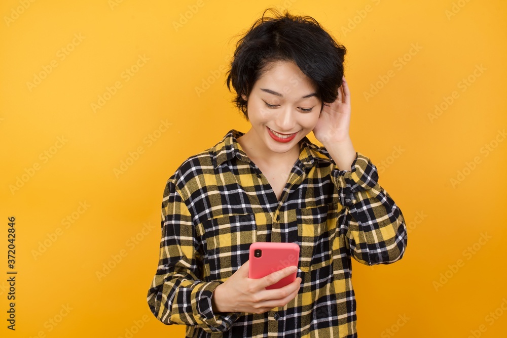 Happy Young beautiful Asian woman wearing plaid shirt over yellow background feels good while listens pleasant melody in headphones, focused in screen of smartphone, People, technology, lifestyle Happy Young beautiful Asian woman wearing plaid shirt over yellow background feels good while listens pleasant melody in headphones, focused in screen of smartphone, People, technology, lifestyle