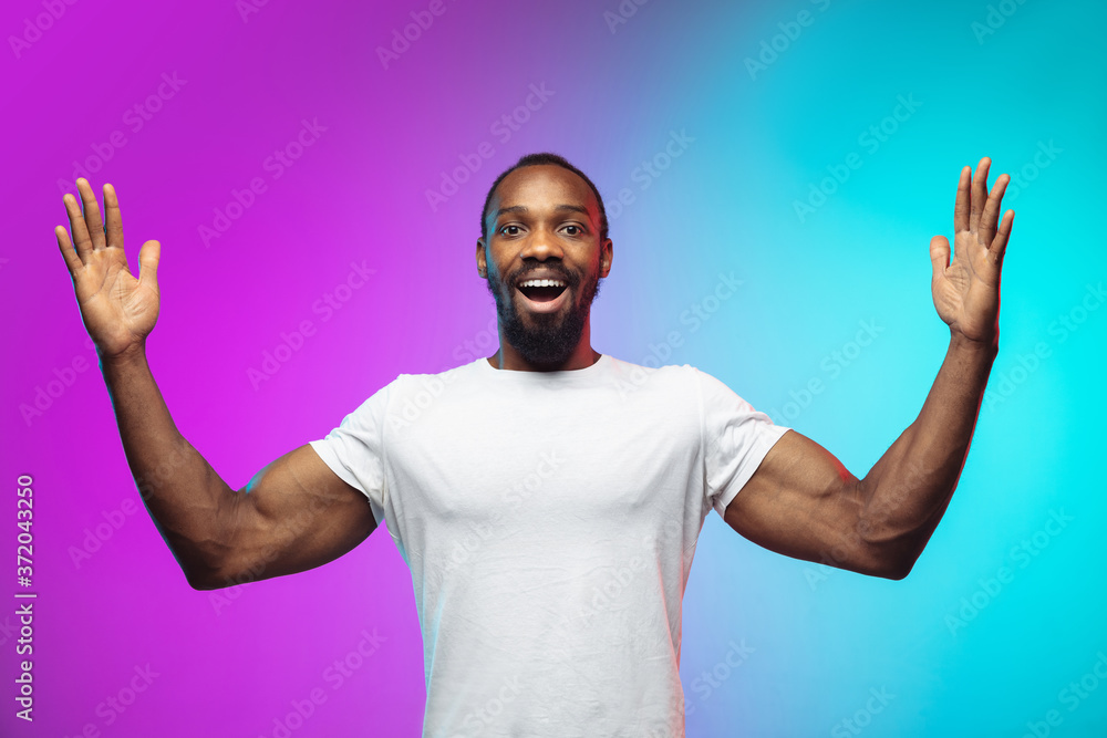 Naklejka premium Showing a huge. African-american young man's portrait on gradient studio background in neon. Beautiful male model in casual style, white shirt. Concept of human emotions, facial expression, sales, ad.