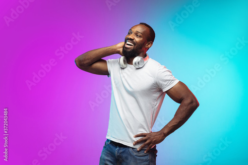 Laughting. African-american young man's portrait on gradient studio background in neon. Beautiful male model in casual style, white shirt. Concept of human emotions, facial expression, sales, ad.