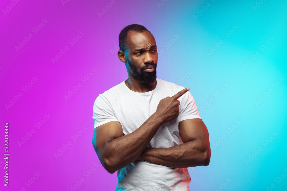 Pointing, showing. African-american young man's portrait on gradient studio background in neon. Beautiful male model in casual style, white shirt. Concept of human emotions, facial expression, sales Pointing, showing. African-american young man's portrait on gradient studio background in neon. Beautiful male model in casual style, white shirt. Concept of human emotions, facial expression, sales