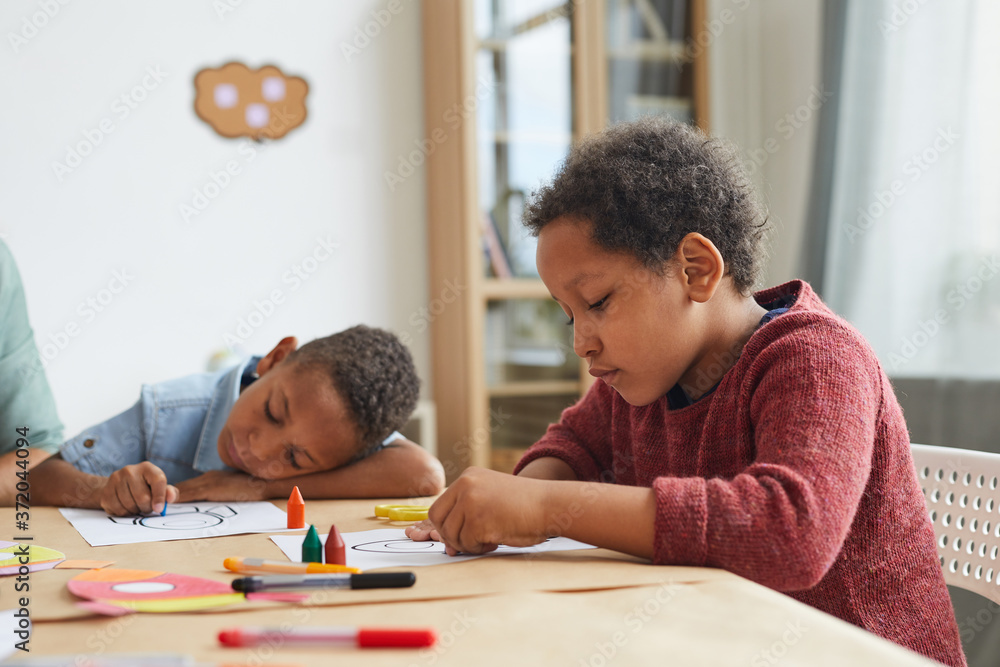 Side view portrait of African-American boy drawing pictures with crayons while enjoying art class in preschool, copy space