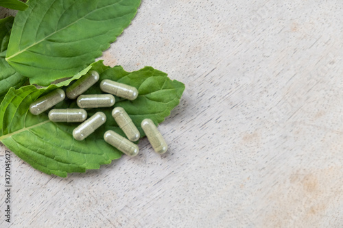 Selective focus at Herbal medicine in capsule on brown wood surface, with green  holy basil leaf herb at the background. Healthcare and medical lifestyle concept.