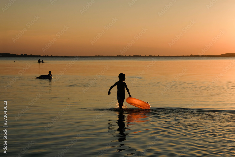 Silhouette of a boy with an orange inflatable circle on a background of sunset. Ukrainian lake Svityaz. Copy space. 