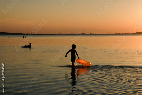 Silhouette of a boy with an orange inflatable circle on a background of sunset. Ukrainian lake Svityaz. Copy space.  © Ganna Zelinska