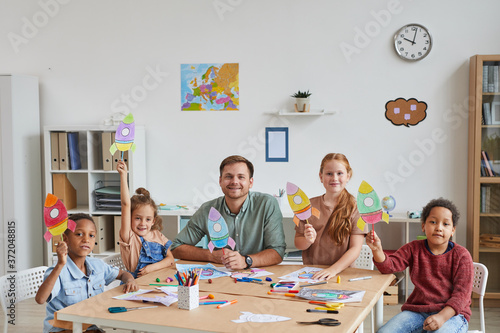 Portrait of smiling male teacher with multi-ethnic group of kids showing pictures of space rockets while enjoying art and craft lesson in preschool or development center, copy space photo