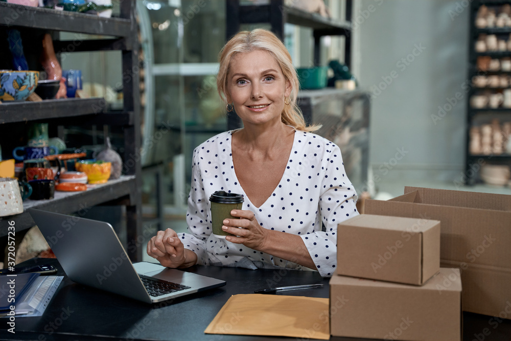 Morning coffee. Portrait of beautiful female business owner or business woman working on laptop in art studio or craft pottery shop, drinking coffee and smiling at camera Morning coffee. Portrait of beautiful female business owner or business woman working on laptop in art studio or craft pottery shop, drinking coffee and smiling at camera