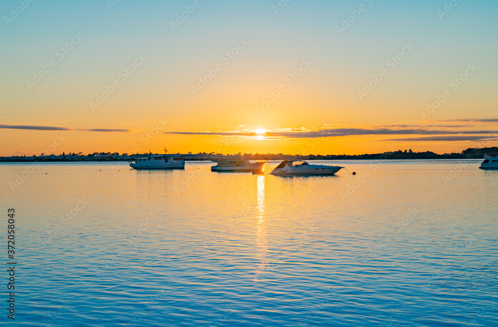 Three boats moored near horizon at sunrise over blue water of Tauranga harbour with intense golden glow on horizon.
