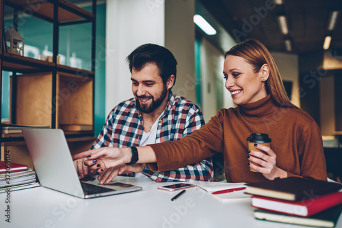 Cheerful internet users laughing and enjoying time after watching webinar online at coworking space, smiling friends sitting at cafeteria and communicated via laptop while girl holding cup coffee