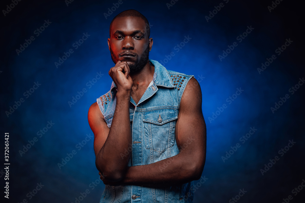 Fototapeta premium Fashionable attractive black guy in jean jacket. An african person with blue foggy background behind. Urban style.