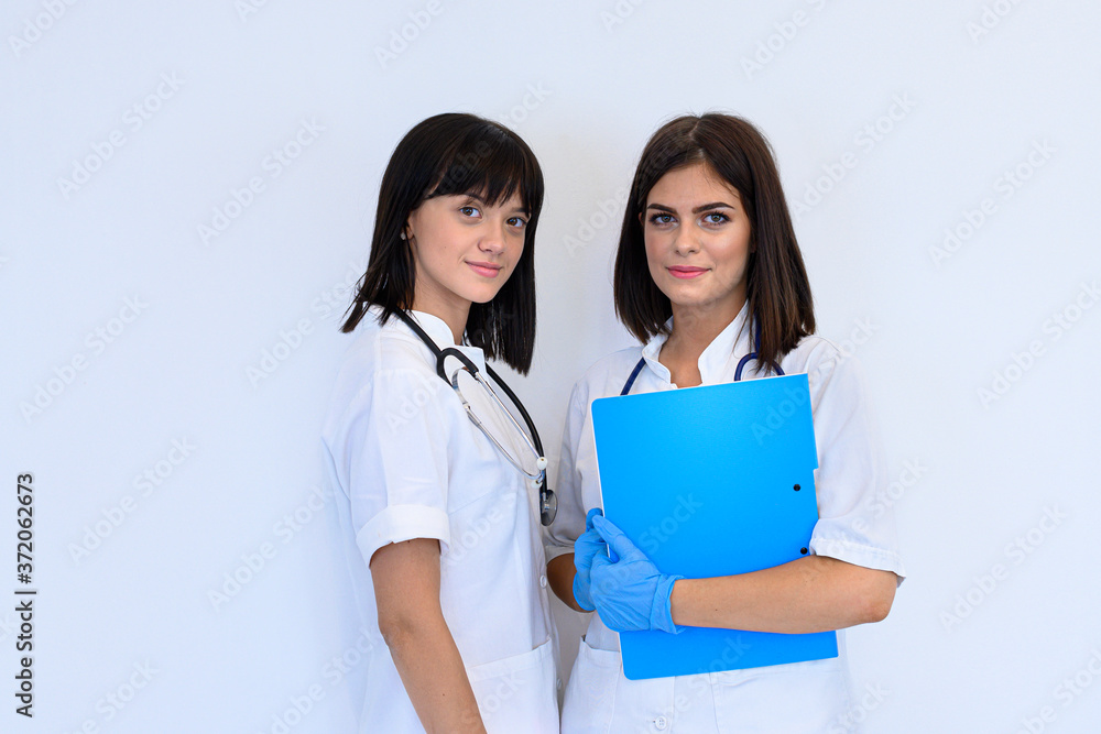 Two young female practitioners in white uniforms with stethoscopes around neck with blue protection gloves. One of them is holding record of temperature, pulse and respiration. Two young female practitioners in white uniforms with stethoscopes around neck with blue protection gloves. One of them is holding record of temperature, pulse and respiration.
