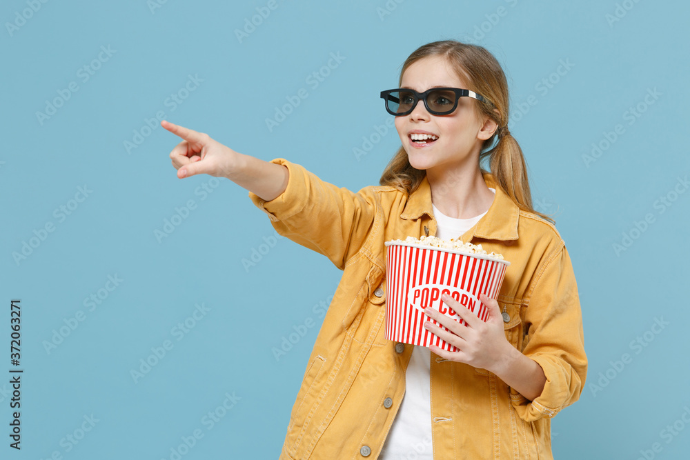 Fototapeta premium Pretty little kid girl 12-13 years old in 3d glasses isolated on blue background. Childhood lifestyle concept. People in cinema. Watching movie film hold bucket of popcorn pointing index finger aside.