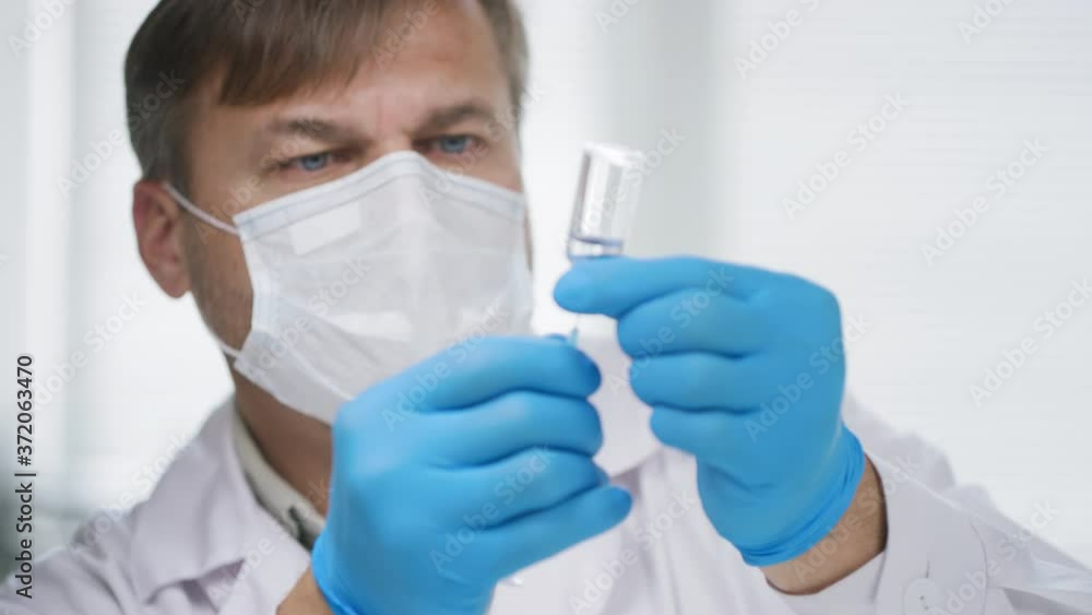 Medium shot of male doctor in face mask and gloves filling syringe with medicine from ampule, then flicking it to remove air bubbles Medium shot of male doctor in face mask and gloves filling syringe with medicine from ampule, then flicking it to remove air bubbles