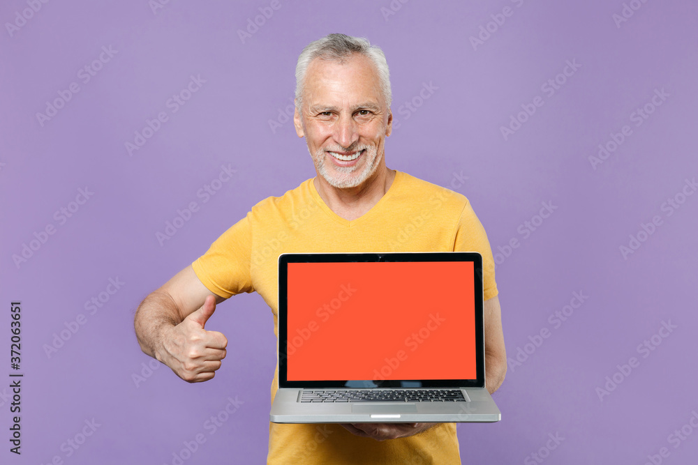 Smiling elderly gray-haired mustache bearded man in casual yellow t-shirt isolated on violet background. People lifestyle concept. Hold laptop pc computer with blank empty screen, showing thumb up. Smiling elderly gray-haired mustache bearded man in casual yellow t-shirt isolated on violet background. People lifestyle concept. Hold laptop pc computer with blank empty screen, showing thumb up.