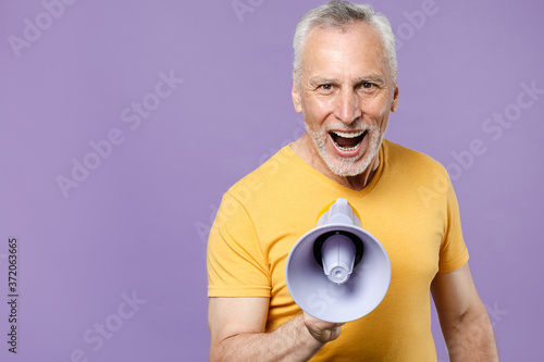 Excited elderly gray-haired mustache bearded man in casual yellow t-shirt posing isolated on violet wall background studio portrait. People sincere emotions lifestyle concept. Screaming in megaphone.