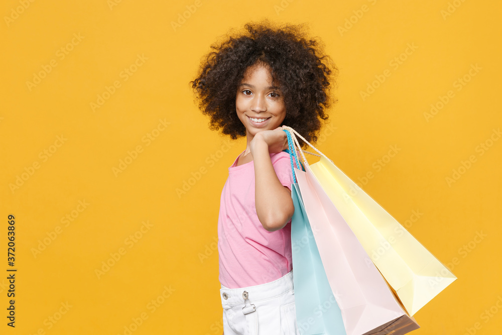 Side view of smiling little african american kid girl 12-13 years old in pink t-shirt isolated on yellow wall background. Childhood lifestyle concept. Hold package bag with purchases after shopping. Side view of smiling little african american kid girl 12-13 years old in pink t-shirt isolated on yellow wall background. Childhood lifestyle concept. Hold package bag with purchases after shopping.