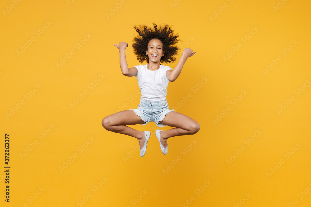 Full length portrait of cheerful little african american kid girl 12-13 years old in white t-shirt isolated on yellow wall background. Childhood lifestyle concept. Jumping, pointing thumbs aside.