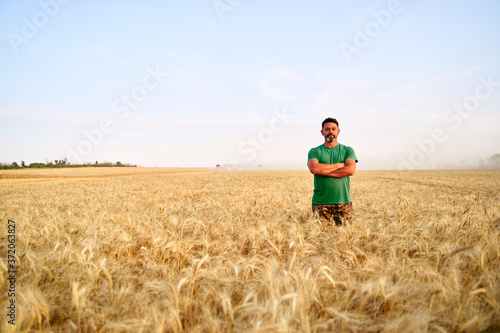 Happy farmer proudly standing in wheat field with arms crossed on chest. Agronomist wearing corporate uniform, looking at camera on farmland. Rich harvest of cultivated cereal crop. Harvesting season. © artiemedvedev