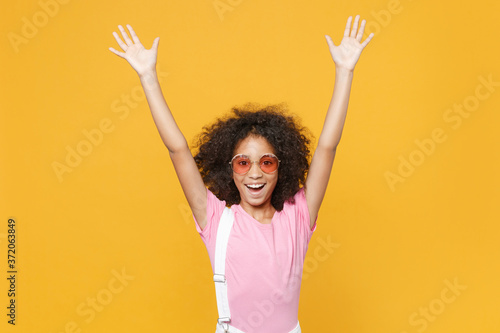 Excited little african american kid girl 12-13 years old in pink t-shirt eyeglasses isolated on yellow wall background studio. Childhood lifestyle concept. Mock up copy space. Rising spreading hands.