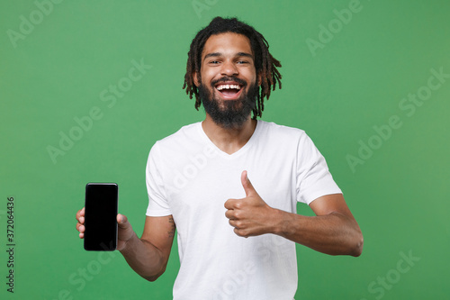 Cheerful young african american man guy 20s in white t-shirt posing hold mobile phone with blank empty screen with mock up copy space showing thumb up isolated on green background studio portrait.