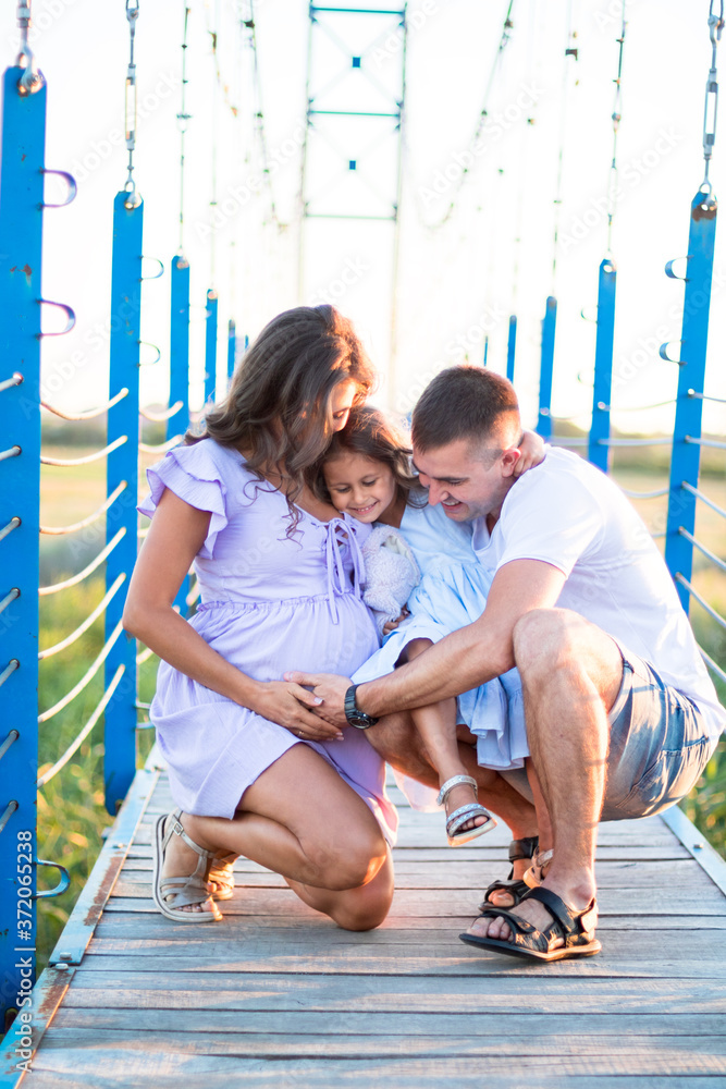Obraz premium happy young family are hugging on the bridge in the sunset light. husband and a little girl touching pregnant wife's belly