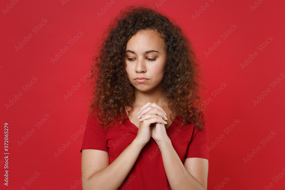 Pensive young african american woman girl in casual t-shirt posing isolated on red background studio portrait. People emotions lifestyle concept. Mock up copy space. Holding hands folded in prayer. Pensive young african american woman girl in casual t-shirt posing isolated on red background studio portrait. People emotions lifestyle concept. Mock up copy space. Holding hands folded in prayer.