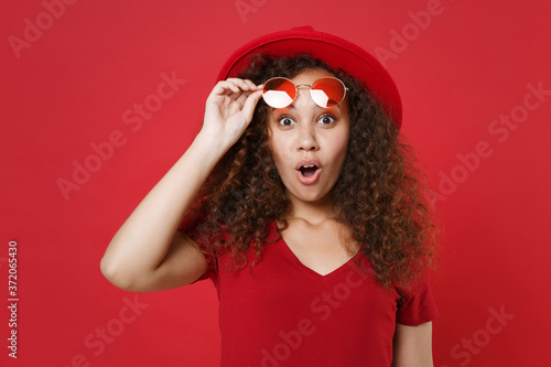 Shocked young african american girl in casual t-shirt hat glasses posing isolated on red background studio portrait. People sincere emotions lifestyle concept. Mock up copy space. Keeping mouth open.