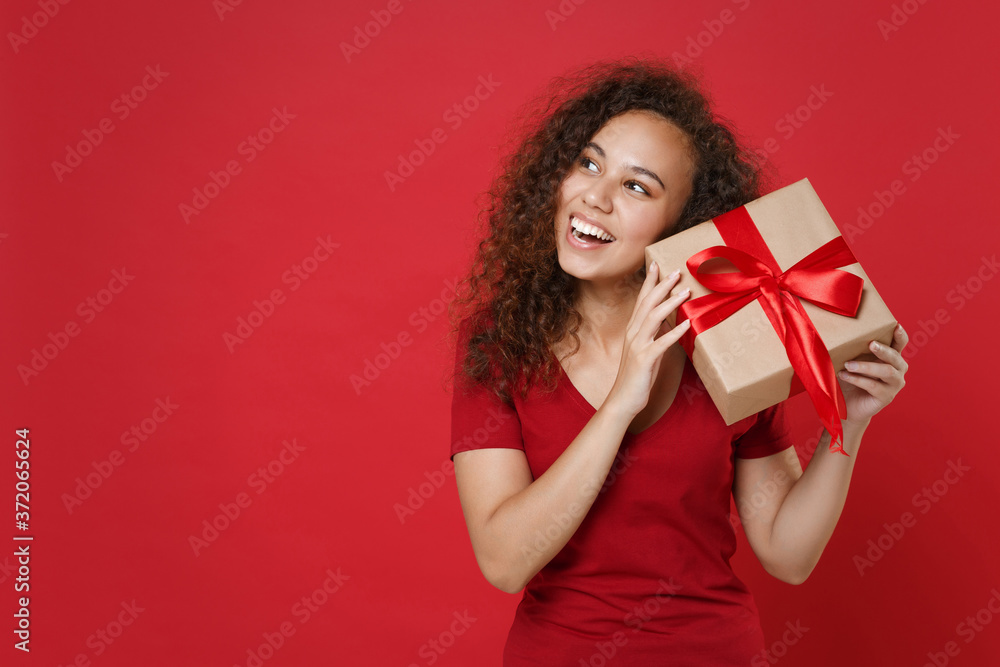 Curious funny young african american woman girl in casual t-shirt posing isolated on red background. St. Valentine's Day, Women's Day, birthday, holiday concept. Hold present box with gift ribbon bow. Curious funny young african american woman girl in casual t-shirt posing isolated on red background. St. Valentine's Day, Women's Day, birthday, holiday concept. Hold present box with gift ribbon bow.