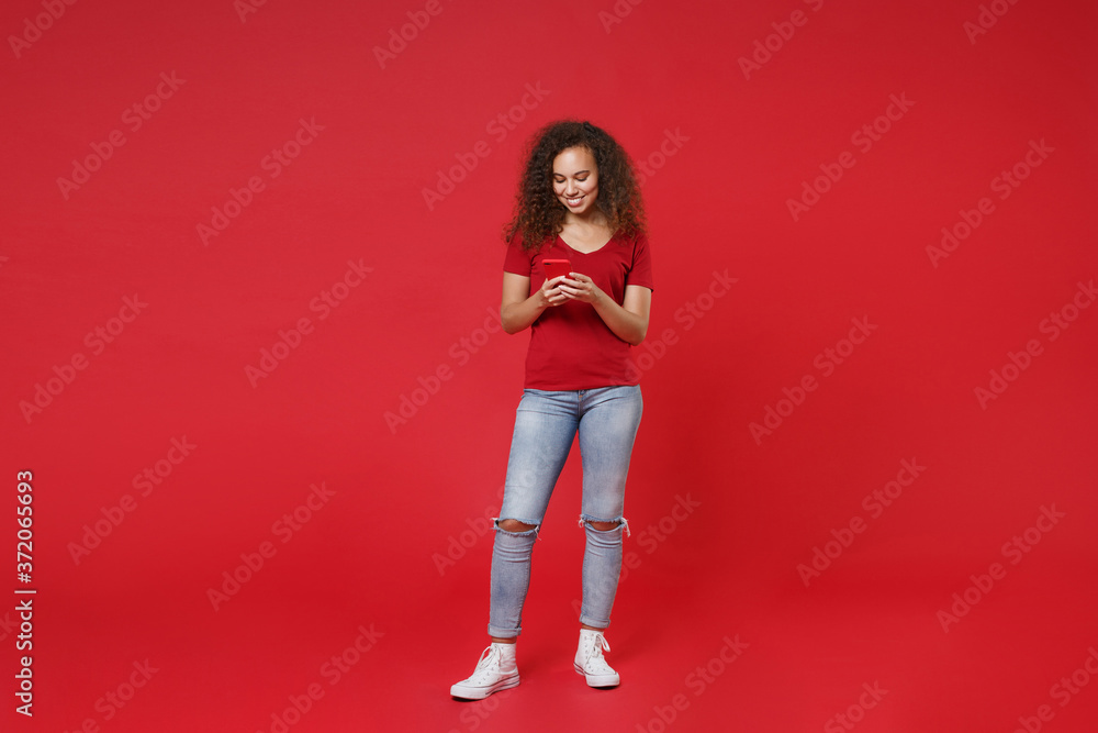 Full length portrait of smiling young african american girl in casual t-shirt isolated on red background. People lifestyle concept. Mock up copy space. Using mobile cell phone, typing sms message. Full length portrait of smiling young african american girl in casual t-shirt isolated on red background. People lifestyle concept. Mock up copy space. Using mobile cell phone, typing sms message.