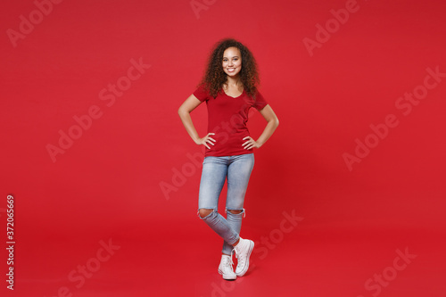 Full length portrait of smiling young african american woman girl in casual t-shirt posing isolated on red background. People lifestyle concept. Mock up copy space. Standing with arms akimbo on waist.