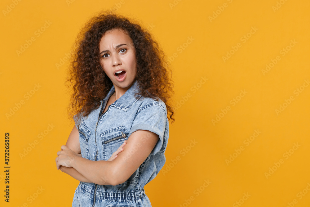 Side view shocked worried young african american woman girl in casual denim clothes isolated on yellow background. People lifestyle concept. Mock up copy space. Holding hands crossed, looking camera. Side view shocked worried young african american woman girl in casual denim clothes isolated on yellow background. People lifestyle concept. Mock up copy space. Holding hands crossed, looking camera.